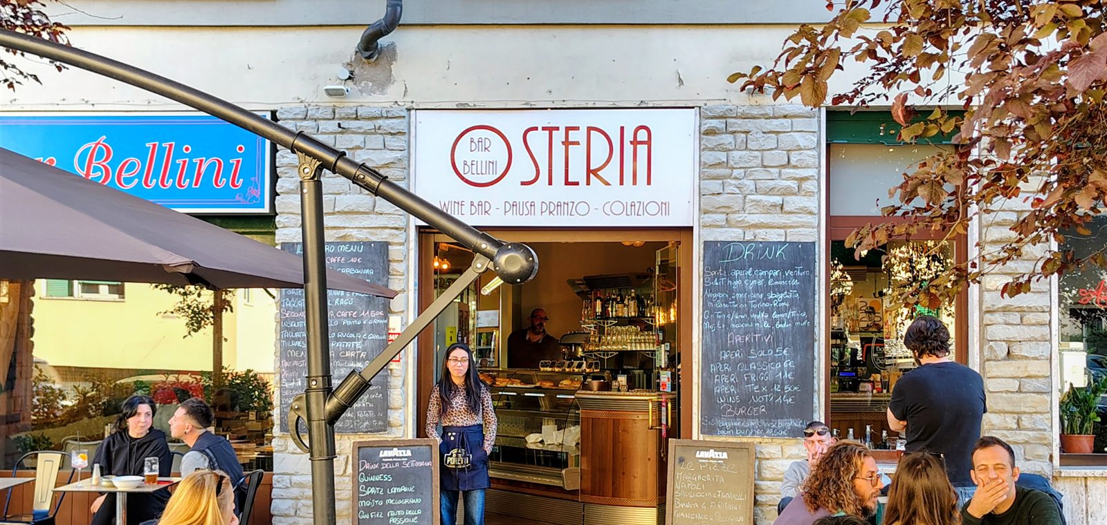 L'esterno del Bar Bellini all'Isolotto: l'insegna Osteria, le lavagne col menu e i tavolini sotto gli alberi